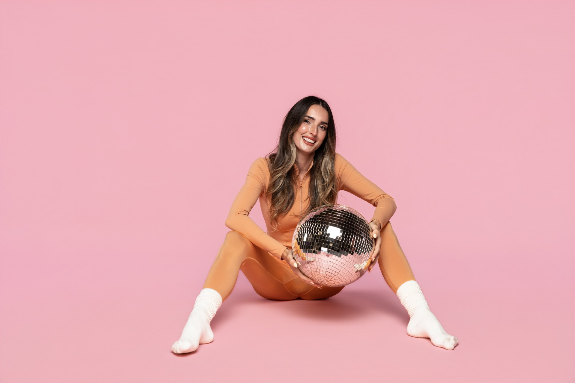 Ash in an orange outfit holding a disco ball against a pink backdrop
